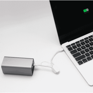 A silver power bank connected to a laptop via a white charging cable. The laptop screen shows a battery charge indicator.