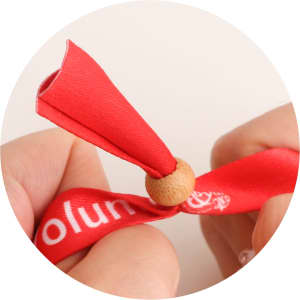 A red RPET fabric wristband with a bamboo toggle, held between two hands. The wristband features a logo.