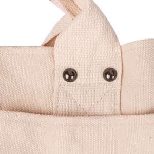 Close-up of a cream canvas tote bag featuring textured fabric and metal studs on the strap attachment.