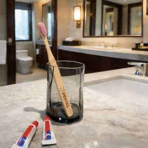 A large flat handle toothbrush with a pink bristle head in a glass, beside two tubes of toothpaste on a bathroom countertop.
