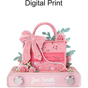 A pink 3D paper art handbag notepad, embellished with flowers and a bow, resting on a flat base with a logo.