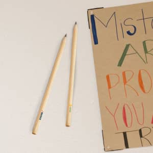 Two wooden grey-lead pencils on a white surface, beside a colourful poster. Each pencil features a logo.