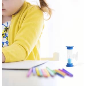 A blue 3-minute sand timer sits beside a child drawing on paper, with colourful pencils scattered in front.