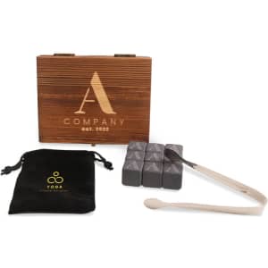 A wooden box with a logo, containing grey whiskey stones, a pair of tongs, and a black velvet pouch.