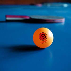An orange table tennis ball with a logo, resting on a blue table, with a paddle in the background.