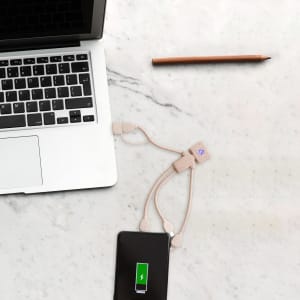 A wheat straw charging cable in a man shape, in beige, connected to a smartphone and a laptop on a marble surface.