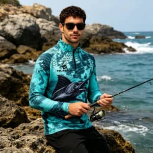 A man wearing a blue and teal fishing pullover with a logo, standing by the water with a fishing rod.