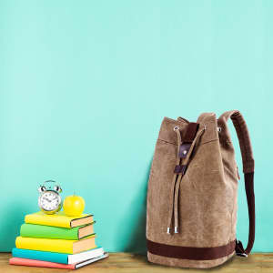 A brown canvas bucket backpack next to a stack of books, an alarm clock, and a green apple on a wooden surface.