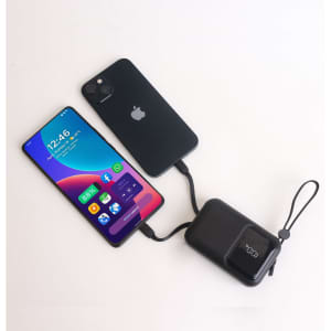 A black portable power bank connected to a smartphone, both displaying charging indicators. The power bank has a logo.