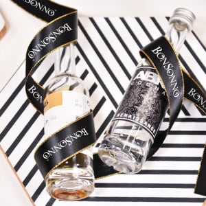 A pair of glass bottles wrapped in a black ribbon with gold edges and a branding logo. The background features a striped design.