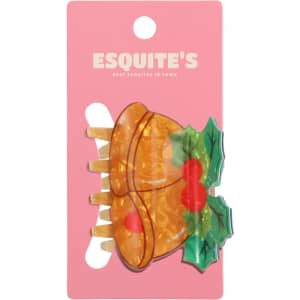 A Christmas-themed hair claw featuring a bell design in orange and green on a pink backing with branding.