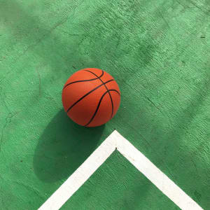 An orange basketball resting on a green court with white lines. The ball has a textured surface and seams.