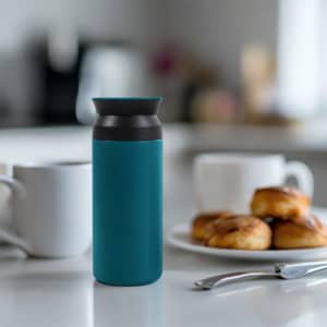 A drink bottle in teal and grey sits on a table, with coffee cups and pastries in the background.