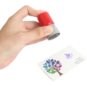 A round stamp with a red top and grey base held above a business card featuring a colourful tree design.