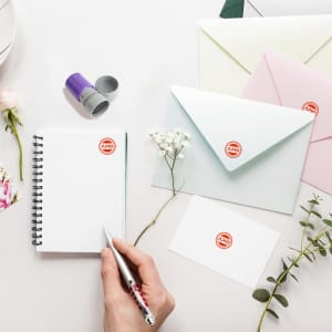 A coloured round stamp in purple, placed alongside envelopes, a notepad, flowers, and a pen on a table.