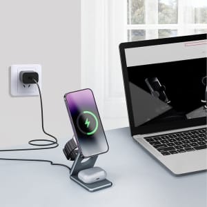 A foldable wireless charger stand in grey, charging a phone and earbuds, placed next to a laptop.