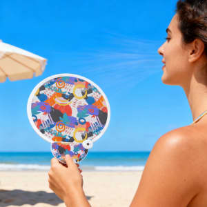 A colourful 20cm foldable fan featuring a vibrant design, held by a person at the beach.