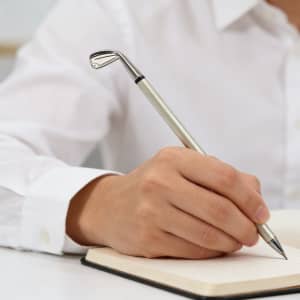 A golf club-shaped pen in silver, held by a person writing in a notebook. The setting features neutral colours.