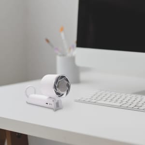 A compact personal cooling fan in white sits on a desk beside a keyboard and a cup with art supplies.
