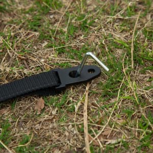 A black strap with a metal peg on grassy ground, designed for securing outdoor equipment.