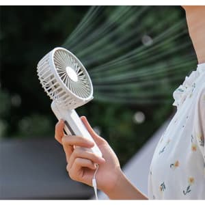 A white foldable USB hand fan is held in a person's hand, with air blowing from its grille. It features a logo.