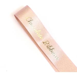 A satin ribbon in a soft pink shade with a metallic gold inscription, featuring a logo.