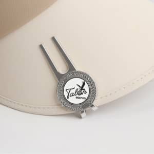 A silver golf divot tool with a ball marker attached, resting on a beige cap. It features a logo design.