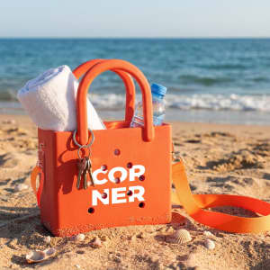 An orange mini beach bag featuring a drink bottle, towel, and keys, resting on sandy beach with waves in the background.