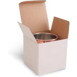 A copper Moscow Mule mug inside an open cardboard box. The mug has a shiny finish and features a logo.