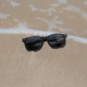 Classic black sunglasses resting on a sandy beach, with gentle waves in the background.