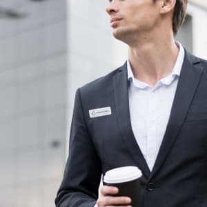 A man in a black suit holding a coffee cup, wearing a custom acrylic name badge on his lapel.