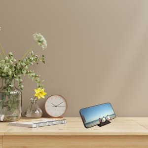 A multi-function phone stand in black, displaying a phone against a neutral background with a clock and flowers nearby.