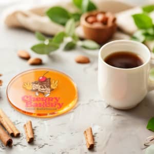 An orange tinted acrylic coaster featuring a logo, beside a white coffee cup and cinnamon sticks.