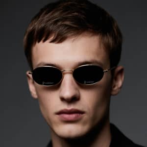 Oval retro sunglasses with a gold frame and dark lenses, worn by a model with short hair against a dark background.
