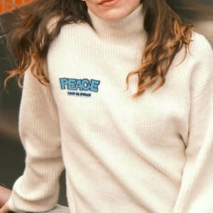 A light cream turtleneck sweater featuring a colourful embroidered patch with the word "PEACE."