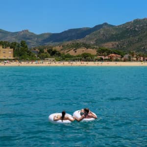 An inflatable swim ring in white floats on clear blue water, with a beach and mountains visible in the background.