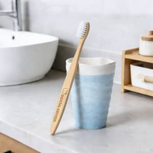 A large bamboo toothbrush with a light wooden handle and white bristles rests against a blue ceramic cup on a countertop.
