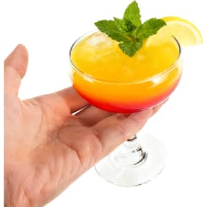 A hand holding a Martini coupe glass filled with a vibrant yellow and red drink, garnished with mint and a lemon slice.