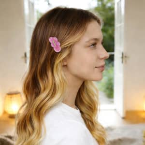 A hair clip featuring a pink flower design, worn in a woman's hair, set against a bright indoor background.
