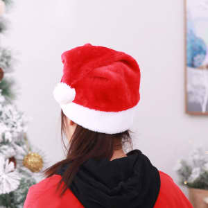 A red plush Santa hat with a white brim and pom-pom, worn on a person's head. Christmas decorations in the background.