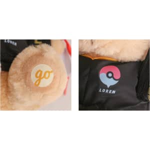 A plush teddy bear with a light brown fur, wearing a graduation outfit and featuring a logo on its ear.
