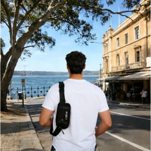 A black travel fanny pack worn across the shoulder of a person standing outdoors by the water, with buildings in the background.
