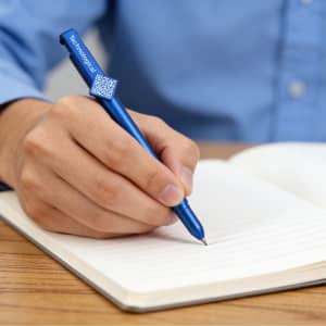A blue pen with a QR code and branding is held above a notebook on a wooden surface.