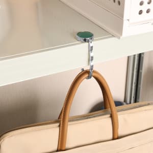 A round foldable desk hook in silver, attached to a shelf, with a tan handbag hanging from it.