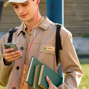 A young man in a beige jacket and cap holds three green books and a smartphone, wearing pink earbuds.