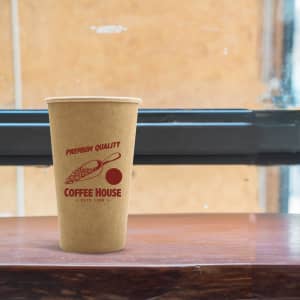 A kraft paper cup in brown, featuring a logo and design, placed on a wooden surface near a window.