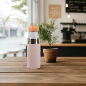 A drink bottle in pastel pink with a silver top and orange lid, placed on a wooden table next to a potted plant.