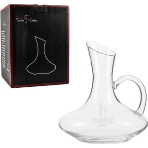 A glass wine decanter with a handle, placed beside its black box featuring a logo. Transparent and elegantly shaped.