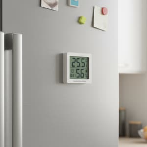 A digital temperature humidity thermometer in white mounted on a silver fridge, displaying temperature and humidity readings.