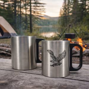 A stainless steel insulated mug with a plastic handle, featuring a design of an eagle. Set outdoors by a campfire.